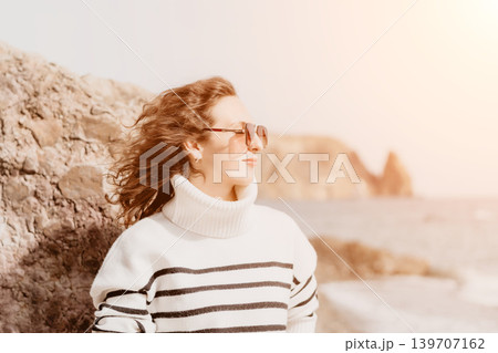 Woman beach vacation standing by rocks in a striped sweater and sunglasses enjoying the coastal scenery Woman beach vacation standing by rocks in a striped sweater and sunglasses enjoying the coastal scenery 139707162
