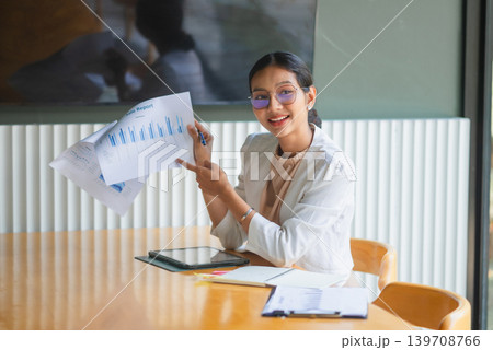 Smiling businesswoman in glasses points to a printed sales report at a wooden meeting table, presenting financial charts with a tablet, laptop, notes, and office documents for teamwork and planning. Smiling businesswoman in glasses points to a printed sales report at a wooden meeting table, presenting financial charts with a tablet, laptop, notes, and office documents for teamwork and planning. 139708766