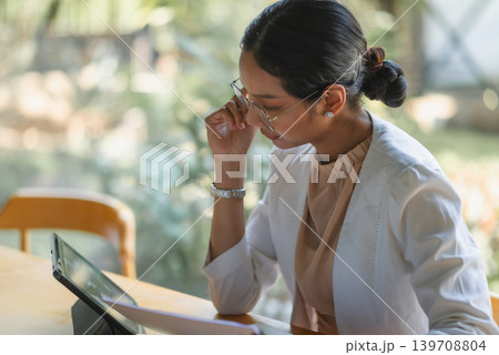 Focused businesswoman in glasses reviews a tablet and documents at a wooden desk, showing financial analysis, digital workflow, research, planning, concentration, and corporate productivity. Focused businesswoman in glasses reviews a tablet and documents at a wooden desk, showing financial analysis, digital workflow, research, planning, concentration, and corporate productivity. 139708804