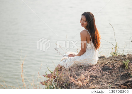 Smiling woman in white lace dress sitting beside calm lake and looking back in warm sunlight, showing relaxed summer vacation, peaceful nature, outdoor wellness, freedom, soft beauty, Smiling woman in white lace dress sitting beside calm lake and looking back in warm sunlight, showing relaxed summer vacation, peaceful nature, outdoor wellness, freedom, soft beauty, 139708806