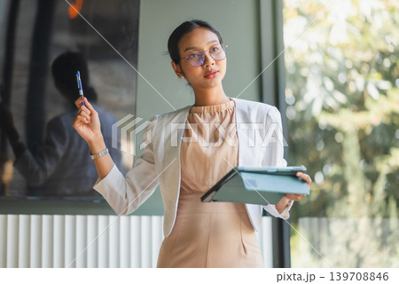 Confident businesswoman in glasses holds a tablet and pen near a meeting screen, representing corporate presentation, leadership, digital planning, teamwork, productivity, and business training. Confident businesswoman in glasses holds a tablet and pen near a meeting screen, representing corporate presentation, leadership, digital planning, teamwork, productivity, and business training. 139708846