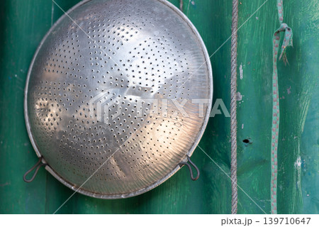 Old metal colander hanging on green wooden wall showcasing rustic kitchen utensil traditional household equipment Old metal colander hanging on green wooden wall showcasing rustic kitchen utensil traditional household equipment 139710647