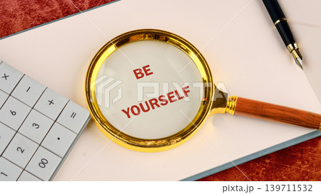 A golden magnifying glass highlights the words BE YOURSELF in red on white paper. A calculator and a dark fountain pen sit on a workspace, suggesting self-reflection A golden magnifying glass highlights the words BE YOURSELF in red on white paper. A calculator and a dark fountain pen sit on a workspace, suggesting self-reflection 139711532