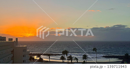 Atlantic ocean sunset with palm trees and volcanic coast in Tenerife Canary Islands. Dramatic evening sky above beach shoreline with travel tourism vacation and tropical leisure. Atlantic ocean sunset with palm trees and volcanic coast in Tenerife Canary Islands. Dramatic evening sky above beach shoreline with travel tourism vacation and tropical leisure. 139711943
