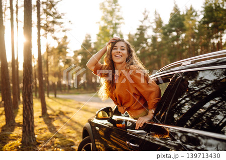 A young woman feels freedom leaning out of a car window in a sunny forest. Travel concept. A young woman feels freedom leaning out of a car window in a sunny forest. Travel concept. 139713430