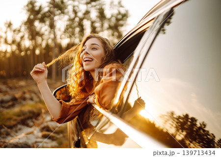A young woman feels freedom leaning out of a car window in a sunny forest. Travel concept. A young woman feels freedom leaning out of a car window in a sunny forest. Travel concept. 139713434