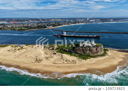 Aerial drone view of Nobbys Lighthouse and Nobbys Beach with a large cargo ship being escorted by tug boats through Newcastle Harbour in the background. Aerial drone view of Nobbys Lighthouse and Nobbys Beach with a large cargo ship being escorted by tug boats through Newcastle Harbour in the background. 139713923
