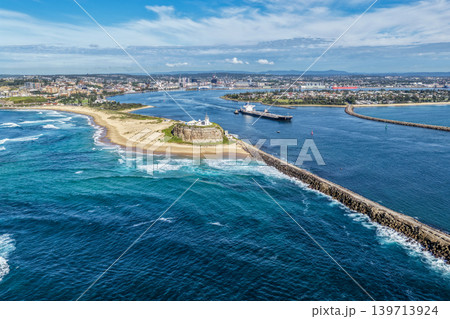 Aerial drone view of Nobbys Lighthouse and Nobbys Beach with a large cargo ship being escorted by tug boats through Newcastle Harbour in the background. 139713924