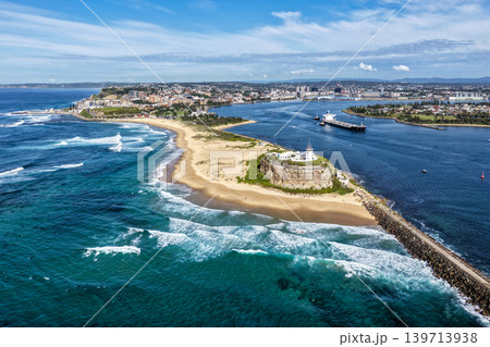 Aerial drone view of Nobbys Lighthouse and Nobbys Beach with a large cargo ship being escorted by tug boats through Newcastle Harbour in the background. Aerial drone view of Nobbys Lighthouse and Nobbys Beach with a large cargo ship being escorted by tug boats through Newcastle Harbour in the background. 139713938