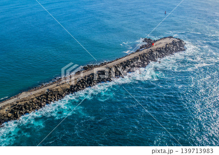 Aerial drone view of the Newcastle breakwall extending into the Pacific Ocean with waves moving along the rocky structure. Aerial drone view of the Newcastle breakwall extending into the Pacific Ocean with waves moving along the rocky structure. 139713983