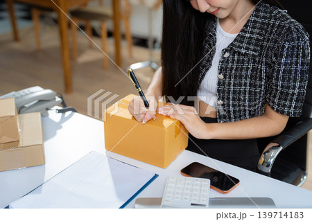 Asian vlogger woman influencer, SME retail store owner writing address on cardboard box at workplace. Asian vlogger woman influencer, SME retail store owner writing address on cardboard box at workplace. 139714183