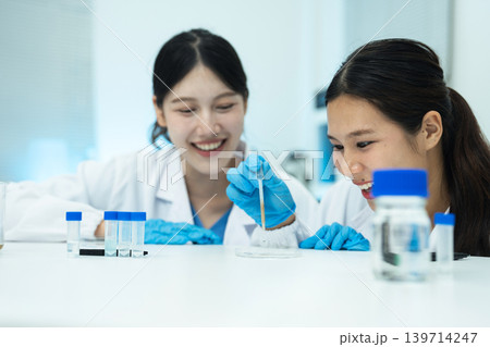 Asian women researchers analyzing cell culture in petri dish near laboratory bottle at workplace. Asian women researchers analyzing cell culture in petri dish near laboratory bottle at workplace. 139714247