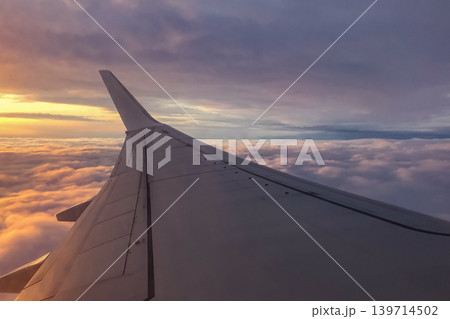 Airplane wing extends above thick clouds during evening flight. Soft sunset light reflects across the sky and aircraft surface. Airplane wing extends above thick clouds during evening flight. Soft sunset light reflects across the sky and aircraft surface. 139714502