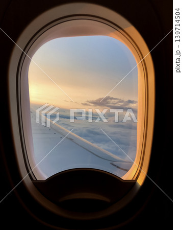 Airplane window frames clouds and wing during a calm evening flight. Soft sunset colors spread across the sky above the horizon. 139714504
