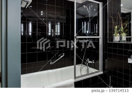 Modern hotel bathroom features black tiles, bathtub, and glass shower screen. Clean fixtures and reflective surfaces create a polished accommodation interior. Modern hotel bathroom features black tiles, bathtub, and glass shower screen. Clean fixtures and reflective surfaces create a polished accommodation interior. 139714513