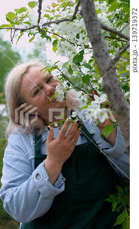 A portrait of a happy woman gardener, standing next to a blossoming apple tree, smiling, enjoying the pleasant scent of the flowers, and feeling happy. A portrait of a happy woman gardener, standing next to a blossoming apple tree, smiling, enjoying the pleasant scent of the flowers, and feeling happy. 139719372