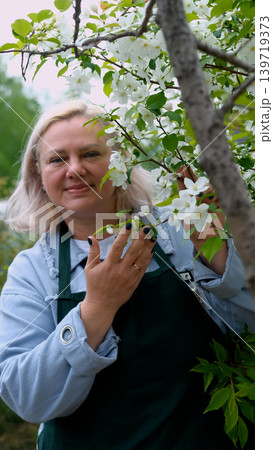 A portrait of a happy woman gardener, standing next to a blossoming apple tree, smiling, enjoying the pleasant scent of the flowers, and feeling happy. A portrait of a happy woman gardener, standing next to a blossoming apple tree, smiling, enjoying the pleasant scent of the flowers, and feeling happy. 139719373