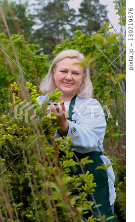 Portrait of happy female gardener standing among bright greenery of her garden Portrait of happy female gardener standing among bright greenery of her garden 139719376