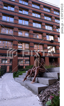 A young beautiful woman with afro-braids, wearing a brown hat, poses while sitting on the steps outside in an eco-friendly residential complex 139719410