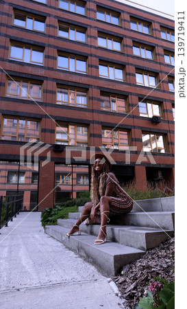 A young beautiful woman with afro-braids, wearing a brown hat, poses while sitting on the steps outside in an eco-friendly residential complex A young beautiful woman with afro-braids, wearing a brown hat, poses while sitting on the steps outside in an eco-friendly residential complex 139719411