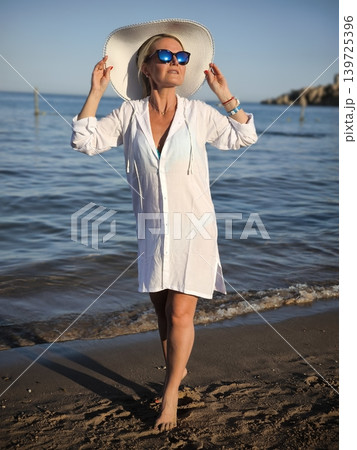 Woman in white tunic and wide-brimmed hat on sandy beach by the sea at sunset Woman in white tunic and wide-brimmed hat on sandy beach by the sea at sunset 139725396