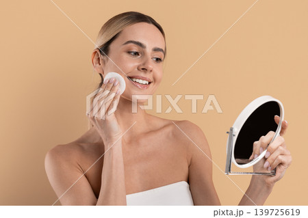 Beautiful smiling woman removing makeup with cotton pad while looking in mirror Beautiful smiling woman removing makeup with cotton pad while looking in mirror 139725619