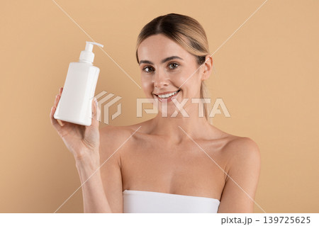 Smiling young woman holding cosmetic lotion bottle and looking at camera Smiling young woman holding cosmetic lotion bottle and looking at camera 139725625