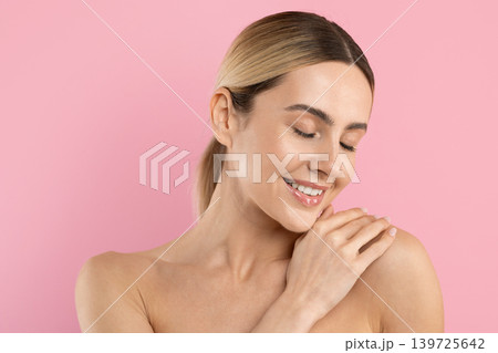 Young woman with radiant skin smiling softly while touching shoulder, pink background Young woman with radiant skin smiling softly while touching shoulder, pink background 139725642