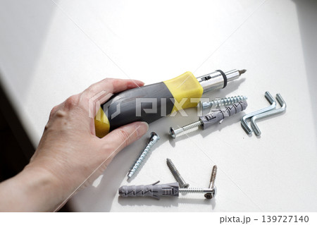 Hand picks up a screwdriver with a black and yellow rubber handle from a white windowsill. Dowels and screws are on the windowsill on a sunny day. 139727140