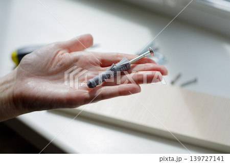 Hand holds a gray plastic dowel against a white windowsill. Dowels and screws are scattered on the windowsill on a sunny day. 139727141