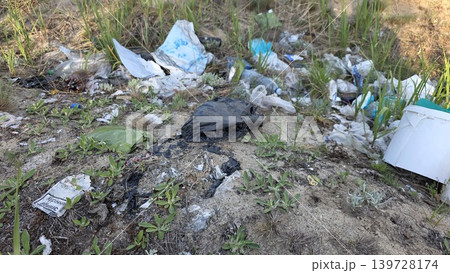 Various garbage including plastic bags, bottles and paper wrappers dumped on a sandy hill with sparse green grass, concept of ecological disaster 139728174