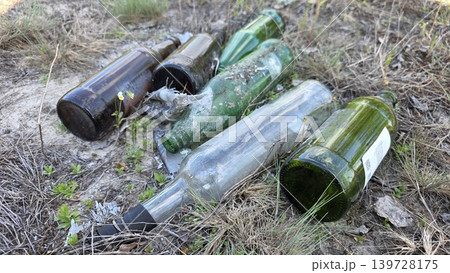 Several empty green and brown glass bottles dumped on sandy ground with dry grass and sparse vegetation close-up, concept of environmental pollution 139728175