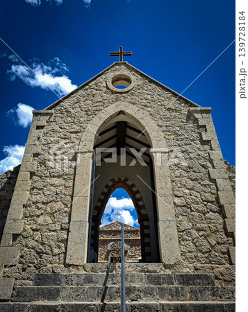A low angle view shows a stone chapel with a cross against a blue sky. The scene is framed by an arched entrance, highlighting the historic design and structure. A low angle view shows a stone chapel with a cross against a blue sky. The scene is framed by an arched entrance, highlighting the historic design and structure. 139728184