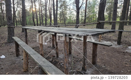 Old rustic wooden picnic table and benches standing on sandy soil in pine forest with scattered garbage. Concept of outdoor recreation area and environmental pollution Old rustic wooden picnic table and benches standing on sandy soil in pine forest with scattered garbage. Concept of outdoor recreation area and environmental pollution 139728203