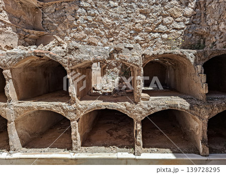 Concrete burial niches stand empty in a ruined catacomb within an old cemetery. Crumbling stone walls surround the area, revealing signs of neglect over time. 139728295
