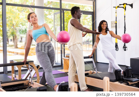 Attentive African American male trainer helping two positive women training with pilates bender ball Attentive African American male trainer helping two positive women training with pilates bender ball 139730564