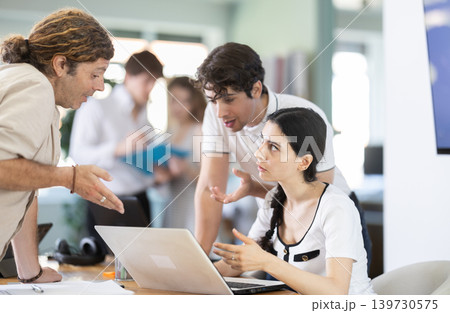 Team businessmen discusses business project while gathered around laptop in company office Team businessmen discusses business project while gathered around laptop in company office 139730575