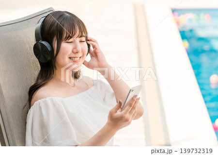 プールサイドのリクライニングチェアに座り音楽を聴く若いアジア人女性 プールサイドのリクライニングチェアに座り音楽を聴く若いアジア人女性 139732319