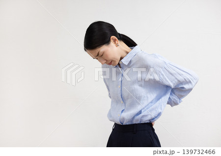 白い壁の前で腰に手を当てて痛そうな表情をする日本人女性 白い壁の前で腰に手を当てて痛そうな表情をする日本人女性 139732466