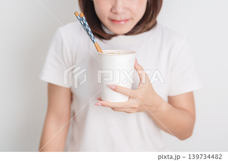 Happy young Asian woman enjoying a delicious cup of hot instant noodles while relaxing at home, Cheerful female eating instant ramen with a smile at apartment. Simple lifestyle and quick meal concept Happy young Asian woman enjoying a delicious cup of hot instant noodles while relaxing at home, Cheerful female eating instant ramen with a smile at apartment. Simple lifestyle and quick meal concept 139734482