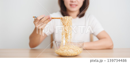 Happy young Asian woman enjoying a delicious bowl of hot instant noodles while relaxing at home, Cheerful female eating instant ramen with a smile at apartment. Simple lifestyle and quick meal concept Happy young Asian woman enjoying a delicious bowl of hot instant noodles while relaxing at home, Cheerful female eating instant ramen with a smile at apartment. Simple lifestyle and quick meal concept 139734486