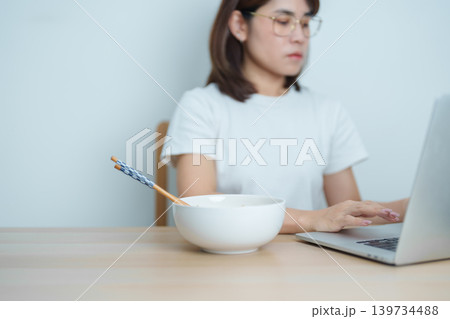 Happy young Asian woman working late at night on laptop while enjoying a bowl of hot instant ramen noodles at home. Cheerful female having a quick meal during work from home. Simple lifestyle concept 139734488