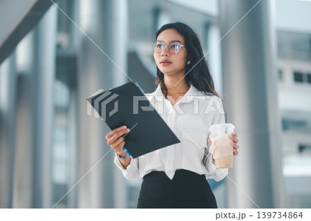 Professional woman in eyeglasses holding iced coffee and a clipboard in a bright office setting, representing calm confidence, corporate lifestyle, waiting time, and everyday work routine. 139734864