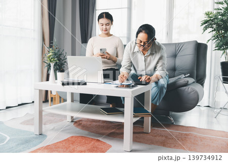 Two women manage household budget with smartphone, laptop, and calculator at home, showing mobile banking, expense planning, bill payment and teamwork in a bright modern living room. Two women manage household budget with smartphone, laptop, and calculator at home, showing mobile banking, expense planning, bill payment and teamwork in a bright modern living room. 139734912
