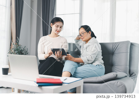 Two women use a tablet while sitting with a laptop at home, discussing budget planning, online consultation, digital learning and teamwork in a bright modern living room workspace. Two women use a tablet while sitting with a laptop at home, discussing budget planning, online consultation, digital learning and teamwork in a bright modern living room workspace. 139734913