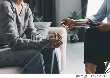 Close up of young woman with clenched hands during mental health counseling session while therapist gestures with notebook, representing anxiety, psychotherapy, support, guidance and recovery. Close up of young woman with clenched hands during mental health counseling session while therapist gestures with notebook, representing anxiety, psychotherapy, support, guidance and recovery. 139734915