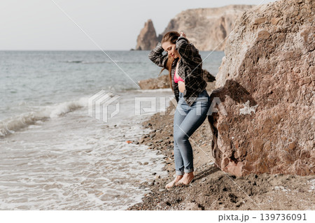Woman beach vacation young woman in jeans and flannel shirt leaning against a large rock on a pebble beach with ocean waves Woman beach vacation young woman in jeans and flannel shirt leaning against a large rock on a pebble beach with ocean waves 139736091