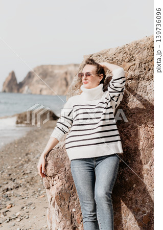 Woman beach coast stylish woman in a striped sweater and sunglasses leans on a rock at a scenic beach with cliffs 139736096