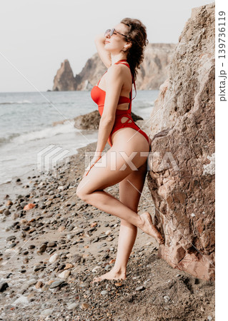 Woman beach swimsuit posing against rocks on a scenic coastline with ocean waves during a relaxing summer vacation Woman beach swimsuit posing against rocks on a scenic coastline with ocean waves during a relaxing summer vacation 139736119