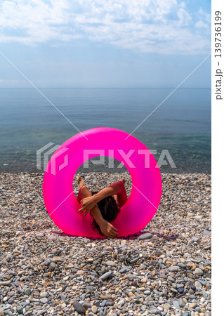 Swim ring relaxation pebble beach, anonymous woman resting inside a neon magenta tube enjoying the summer seascape view 139736199